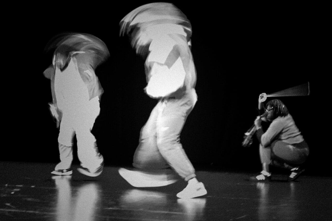 Three people on a dark stage; two are standing and appear blurred by motion, whilst one person is crouching to the right, holding an object near their face. The high-contrast black-and-white image captures the essence of Dance Horizons and dance innovations.