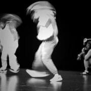 Three people on a dark stage; two are standing and appear blurred by motion, whilst one person is crouching to the right, holding an object near their face. The high-contrast black-and-white image captures the essence of Dance Horizons and dance innovations.