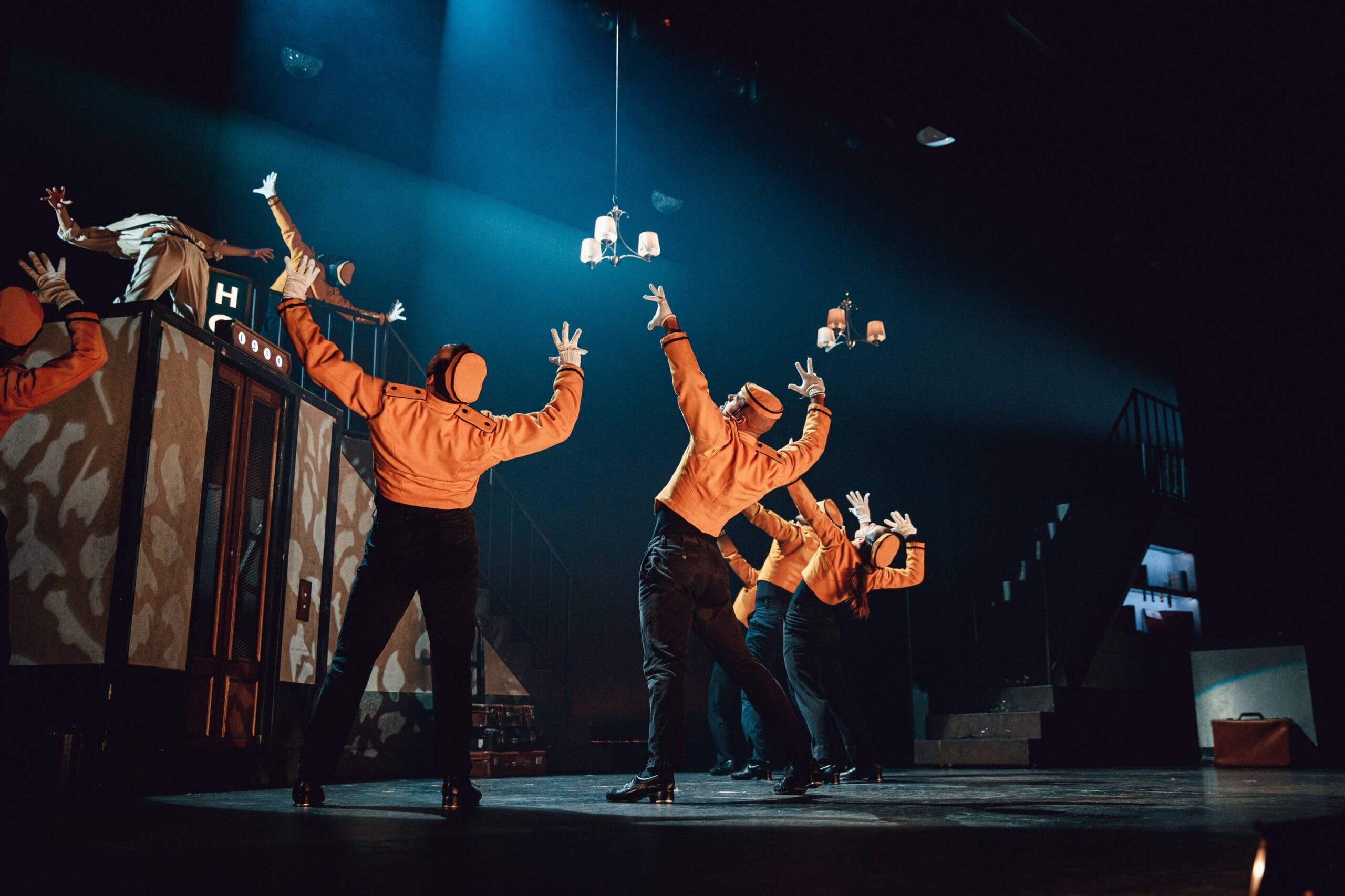 Six performers in orange jackets and black trousers deliver a Tap Dance Sensation, energetically dancing on stage with arms raised. A set resembling a vintage lift and stairs sits in the background under dramatic stage lighting.