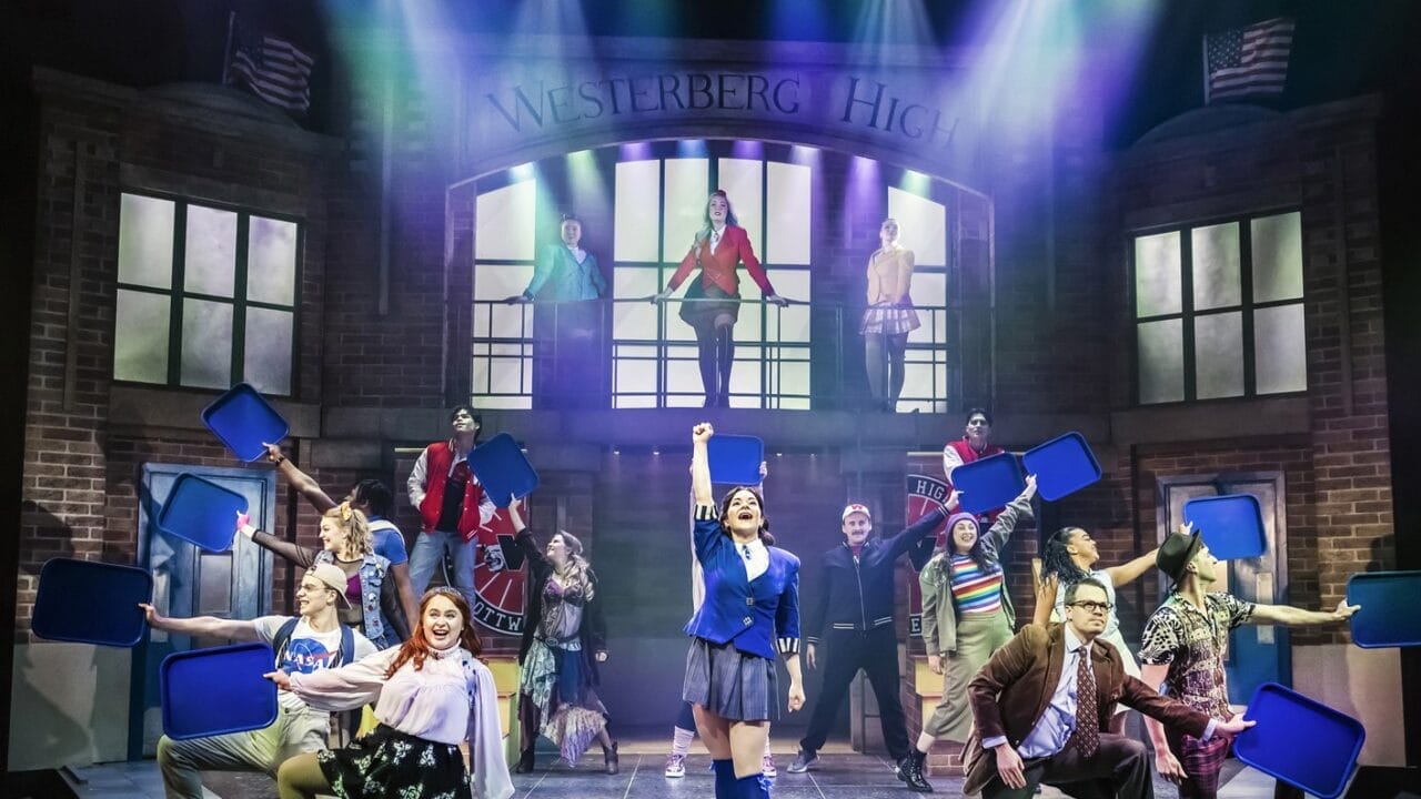 A group of actors perform on stage in front of a set designed as a secondary school entrance labelled "Westerberg High." Some actors hold blue props, and coloured lights shine down, suggesting a musical or theatre production.
