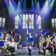 A group of actors perform on stage in front of a set designed as a secondary school entrance labelled "Westerberg High." Some actors hold blue props, and coloured lights shine down, suggesting a musical or theatre production.