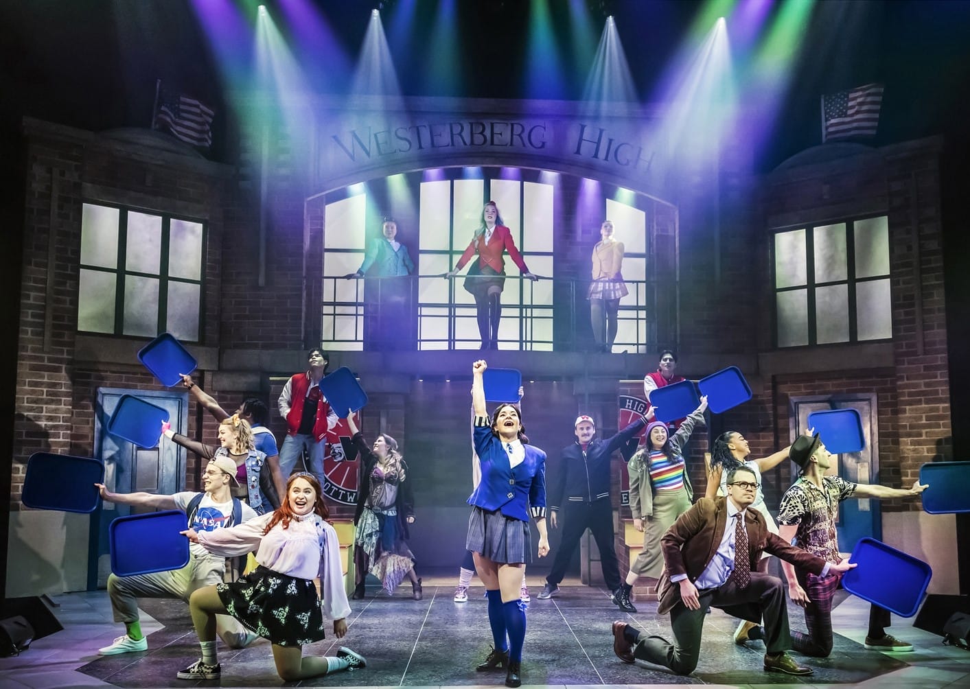 A group of actors perform on stage in front of a set designed as a secondary school entrance labelled "Westerberg High." Some actors hold blue props, and coloured lights shine down, suggesting a musical or theatre production.