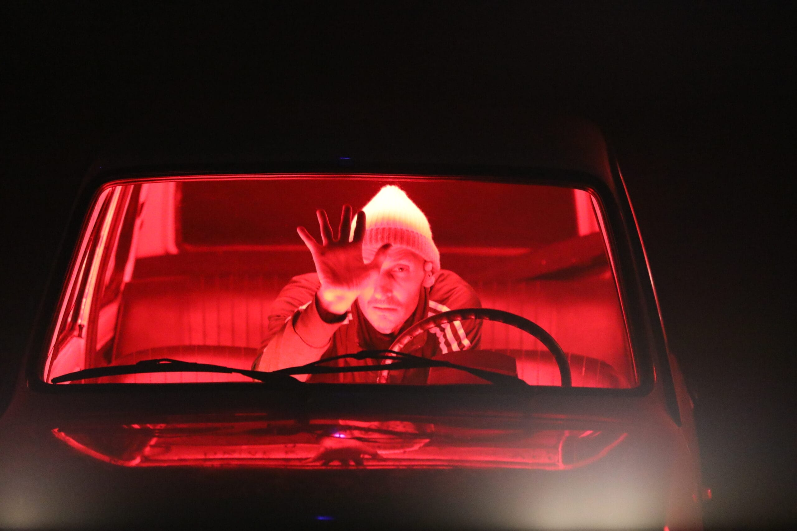 A person wearing a white beanie sits behind the wheel of a car, illuminated by red light, reaching out with one hand towards the windscreen.