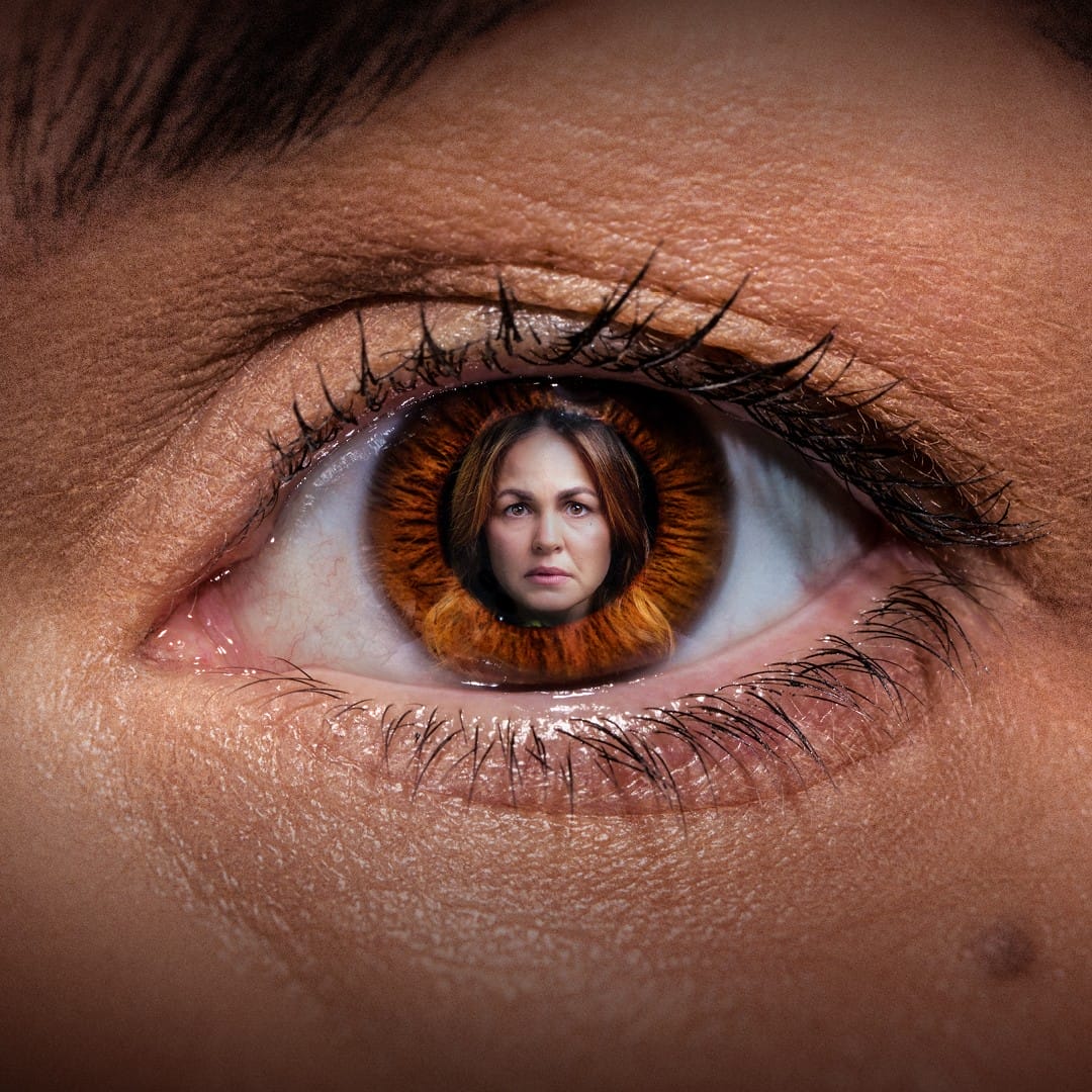A close-up of a human eye with a brown iris, featuring the reflection of a woman’s face centred within the pupil. The eyebrow and eyelashes are clearly visible.