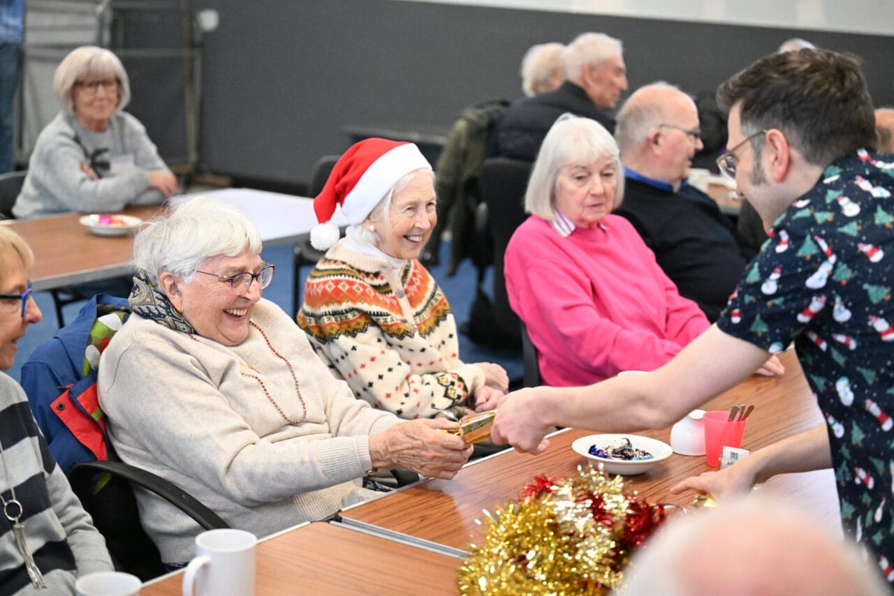 A group of older adults sit at a table in a Care Home, some smiling and one wearing a Santa hat. A person hands a small plate of chocolates across the table, which is decorated with cups, tinsel, and festive decorations.