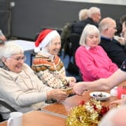 A group of older adults sit at a table in a Care Home, some smiling and one wearing a Santa hat. A person hands a small plate of chocolates across the table, which is decorated with cups, tinsel, and festive decorations.