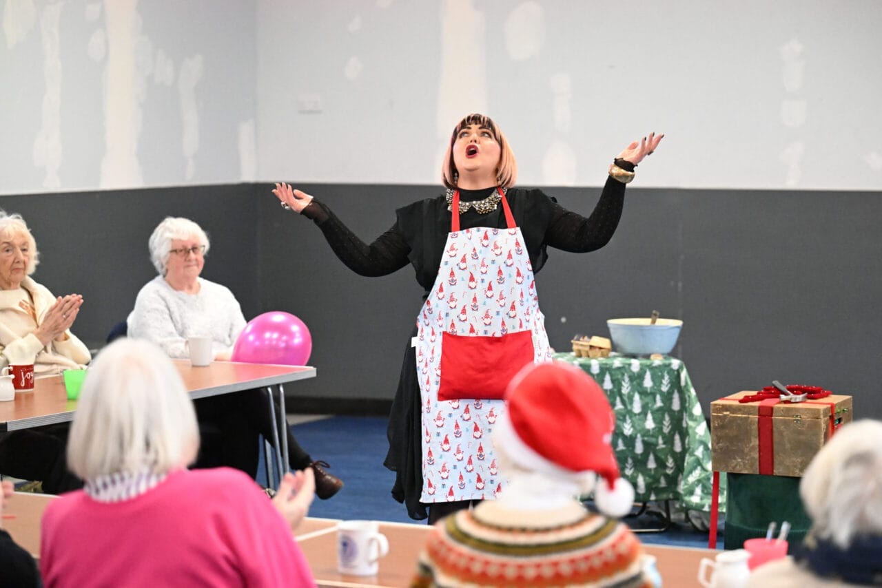 A person in a festive apron, leading Care Home Tours, gestures dramatically between tables of seated people—including some elderly guests. Mugs, a pink balloon, and wrapped presents add to the cheerful atmosphere in the room.