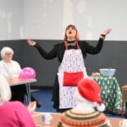 A person in a festive apron, leading Care Home Tours, gestures dramatically between tables of seated people—including some elderly guests. Mugs, a pink balloon, and wrapped presents add to the cheerful atmosphere in the room.