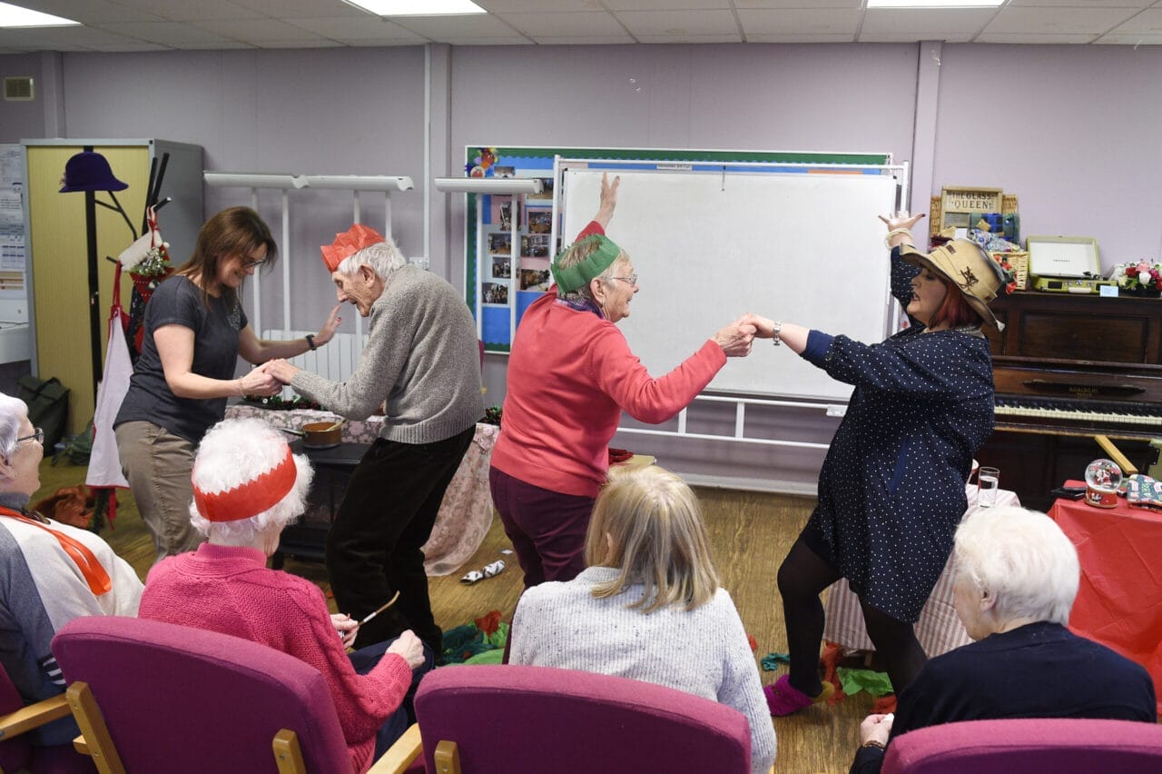 A group of elderly people and two younger women take part in a festive activity during Care Home Tours. Some are seated in pink chairs, while others stand and dance amid holiday hats and cheerful decorations in the communal room.