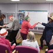 A group of elderly people and two younger women take part in a festive activity during Care Home Tours. Some are seated in pink chairs, while others stand and dance amid holiday hats and cheerful decorations in the communal room.