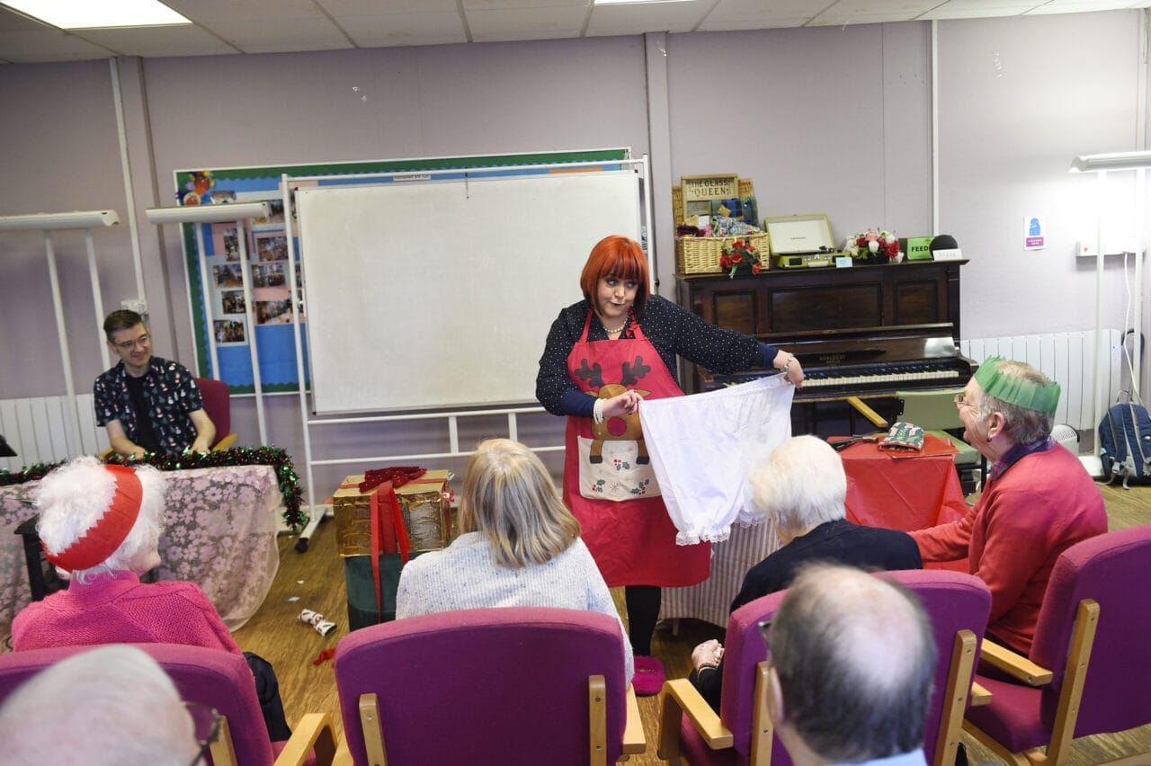 During Care Home Tours, a woman with red hair and an apron stands before seated elderly people, holding up a pair of knickers. A man sits at a table on the left, with a piano and whiteboard in the background.