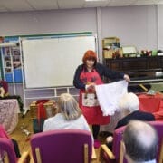 During Care Home Tours, a woman with red hair and an apron stands before seated elderly people, holding up a pair of knickers. A man sits at a table on the left, with a piano and whiteboard in the background.