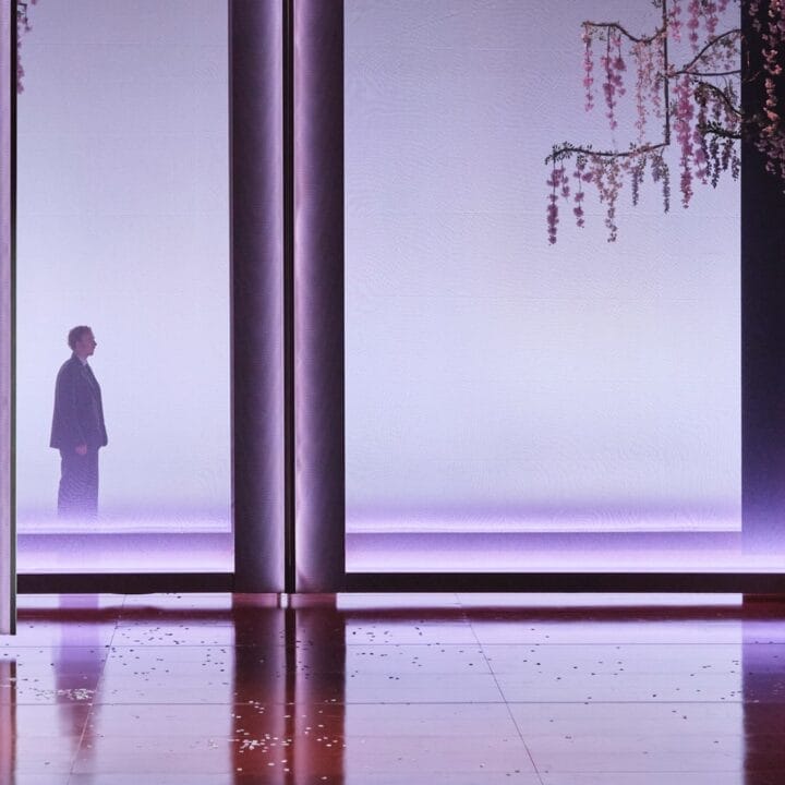 A man in a white naval uniform stands on stage under purple lighting. Cherry blossoms hang from above, evoking the iconic set design of the opera Madama Butterfly.