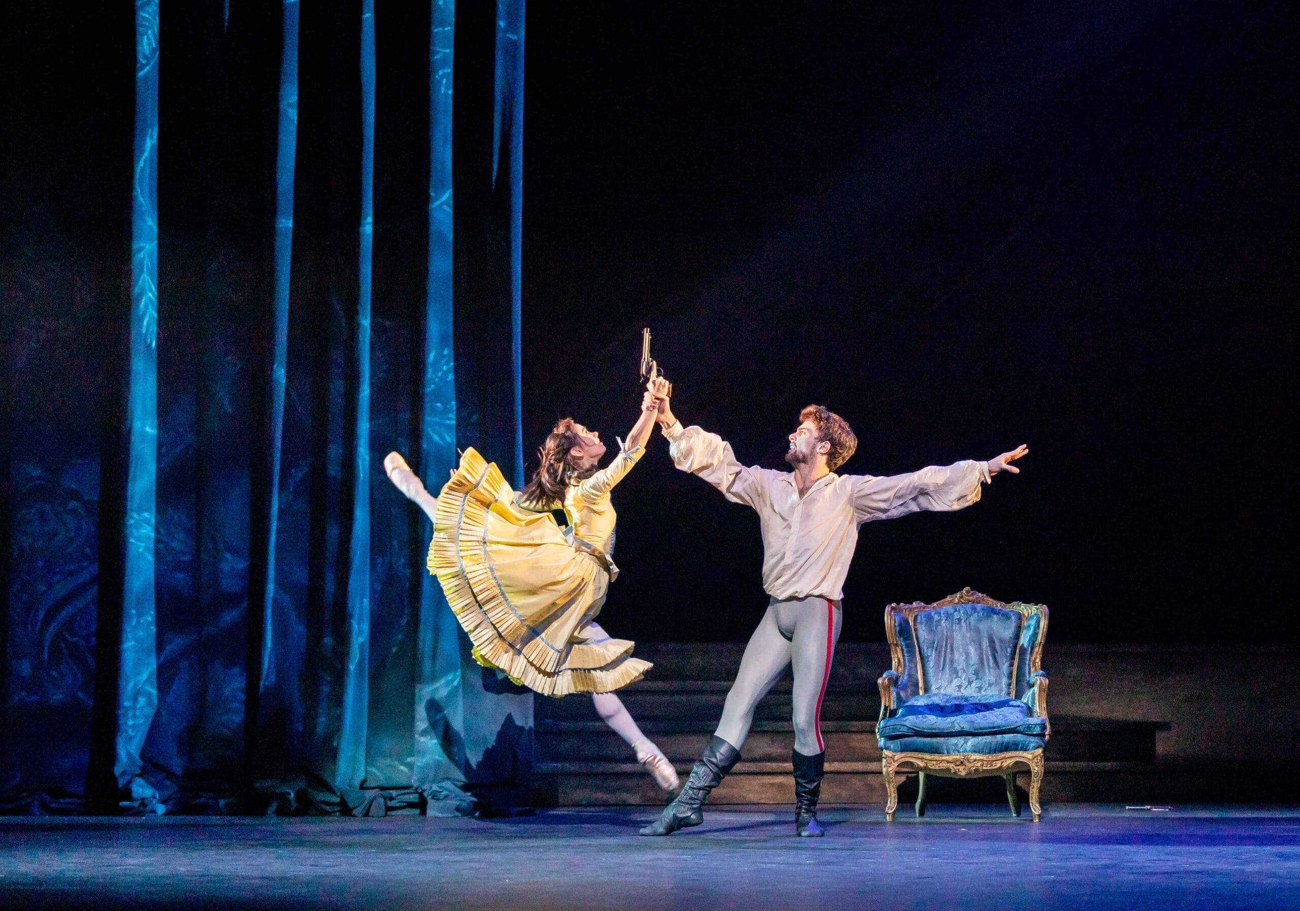 Two ballet dancers perform on stage. The female dancer, in a yellow dress. The male dancer extends his arm, while a blue chair sits nearby.